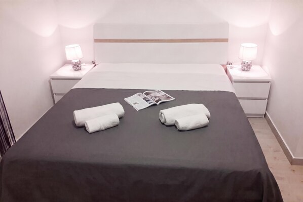 Un lit bien fait, recouvert d’une couverture sombre, avec deux serviettes blanches et un magazine posé sur le lit.