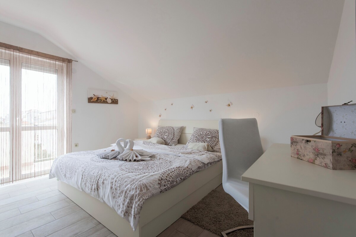 A bedroom with a bed, a desk, a chair, a window with curtains, and a wall-mounted light fixture.