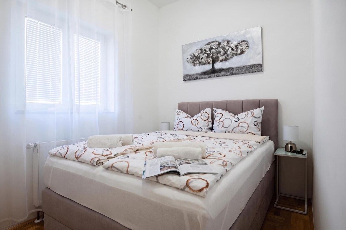 A bedroom with a bed, a bedside table, a lamp, and a window with blinds.