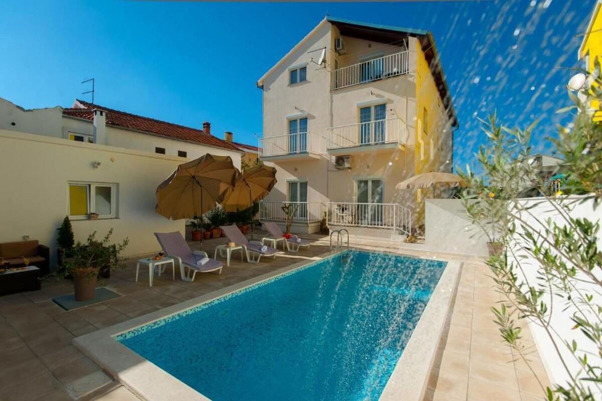A swimming pool with lounge chairs and umbrellas, a multi-story building with balconies, and a clear blue sky.
