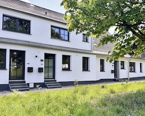 Ein zweistöckiges Gebäude mit weißer Fassade, schwarzen Fensterrahmen und einer schwarzen Tür. Vor dem Eingang führen Stufen hinauf, und im Vordergrund ist ein Baum teilweise sichtbar.