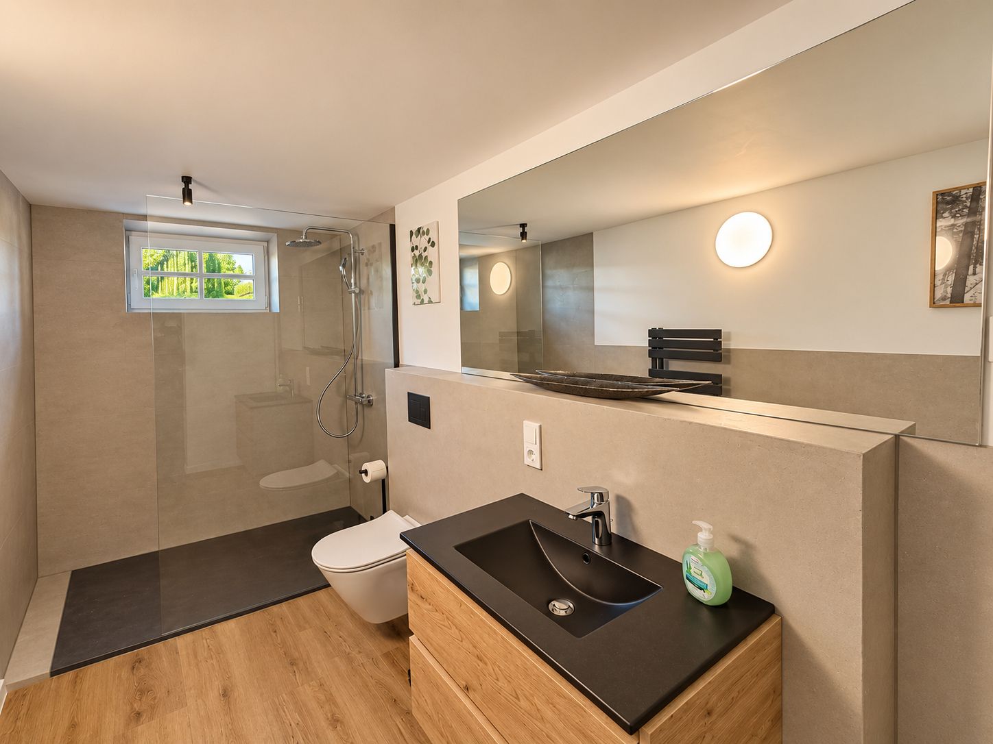 Ein modernes Badezimmer mit einer gläsernen Duschkabine, einer weißen Toilette, einem schwarzen Waschbecken mit Holzunterschrank und einem großen Spiegel mit runder Beleuchtung.