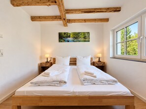 A double bed with white linens, wooden headboard, two bedside tables with lamps, a painting of a landscape, and a window with a view of trees. - Holiday apartment Bergblick in Winterberg for 4 guests (Winterberg)