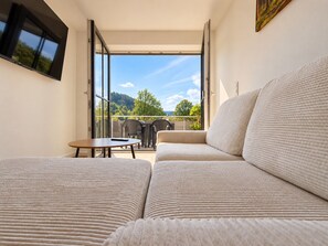 Ein Balkon mit einer Couch und einem Tisch, einem an der Wand montierten Fernseher und Blick auf Bäume und Hügel.