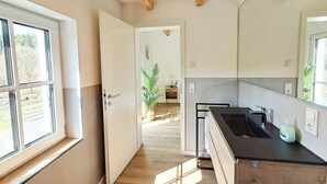 Ein schmaler Flur mit einer zur Kammer führenden Tür, einem Fenster mit Aussicht und einer Küchenzeile mit Spüle und Arbeitsfläche.