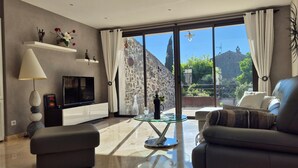 A modern living room with a sofa, TV, and glass table, a balcony with a stone wall, and a clear view of the outside. - Villa Dans le Golfe de Saint-tropez (Cogolin)
