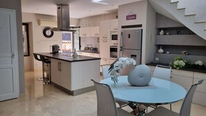 A modern kitchen with a central island, stainless steel appliances, and a dining area with a round table and chairs. - Villa Dans le Golfe de Saint-tropez (Cogolin)