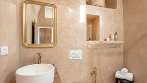 A bathroom with a large oval sink, a mirror with a gold frame, a towel rack, and a shelf with toiletries. - Casa Adamare - Charming Luxury Stay (Ostuni)