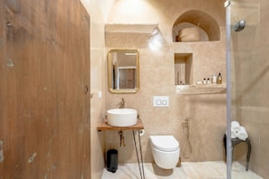 Un baño con tocador de madera, un lavamanos blanco, un grifo dorado, un inodoro blanco, una ducha con puerta de vidrio y un perchero con toallas.