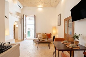 Ein Wohnzimmer mit einer Couch, einem Couchtisch und einem Fernseher. Es gibt einen Esstisch mit Untersetzern und einer Topfpflanze.