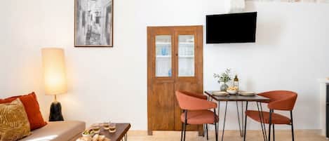 A living room with a sofa, a table with a lamp, a wooden door, and a TV mounted on the wall.