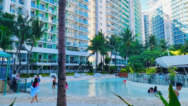 A resort with high-rise buildings, a swimming pool, and palm trees. - Soothing 2-bedrooms lodge with Perfect manmade beach & pool view (Parañaque)