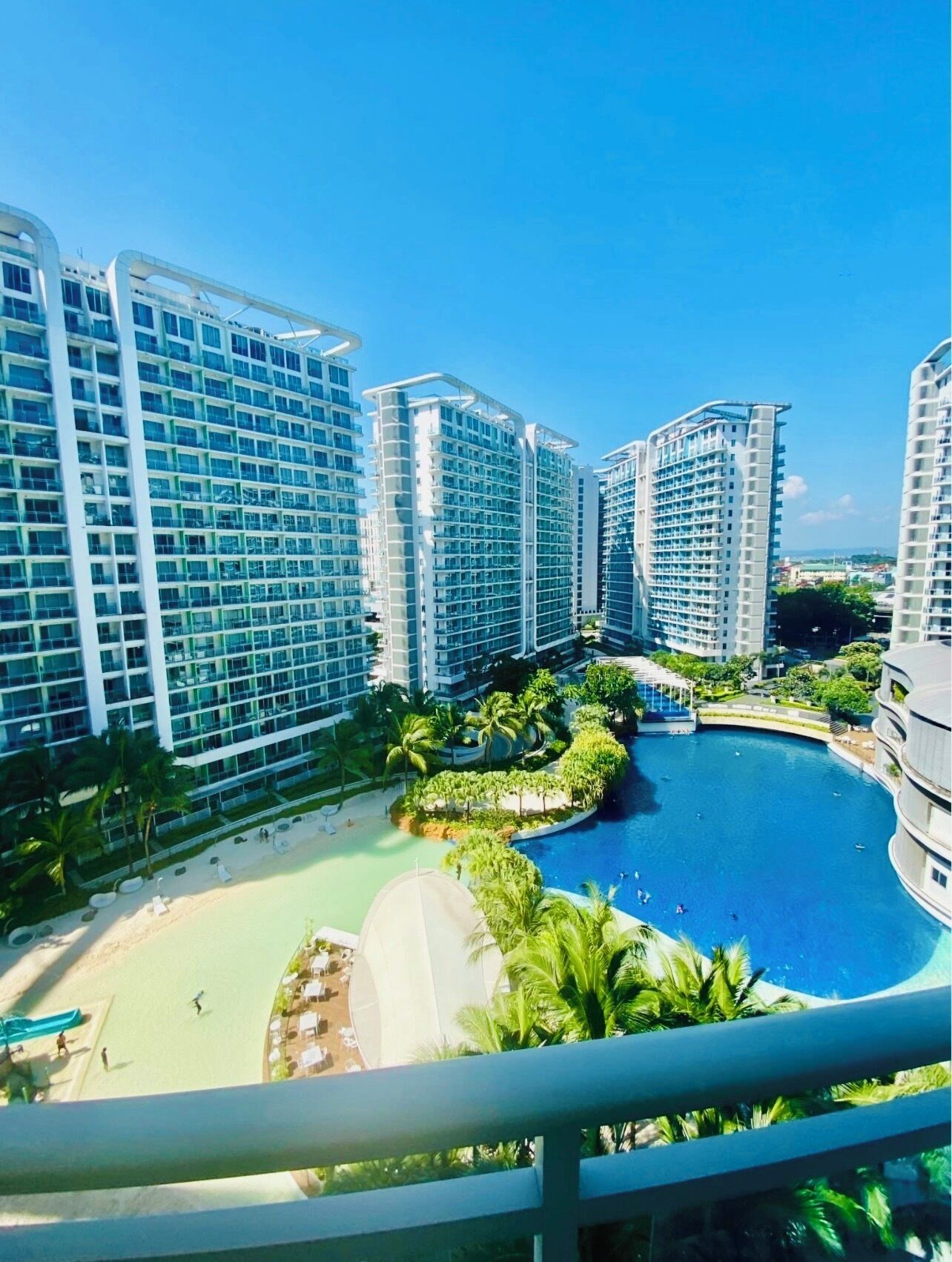 A high-rise hotel with multiple balconies, a central swimming pool, and a landscaped area with palm trees.