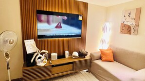 A living room with a flat-screen TV mounted on a wooden panel wall, a white sofa, a small coffee table, a fan, and a decorative light. - Soothing 2-bedrooms lodge with Perfect manmade beach & pool view (Parañaque)