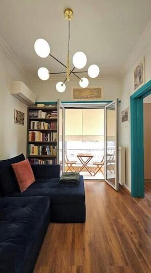 Ein Wohnzimmer mit einem dunkelblauen Sofa, einem Bücherregal, einer Deckenleuchte und Blick durch eine Glastür auf einen Balkon.