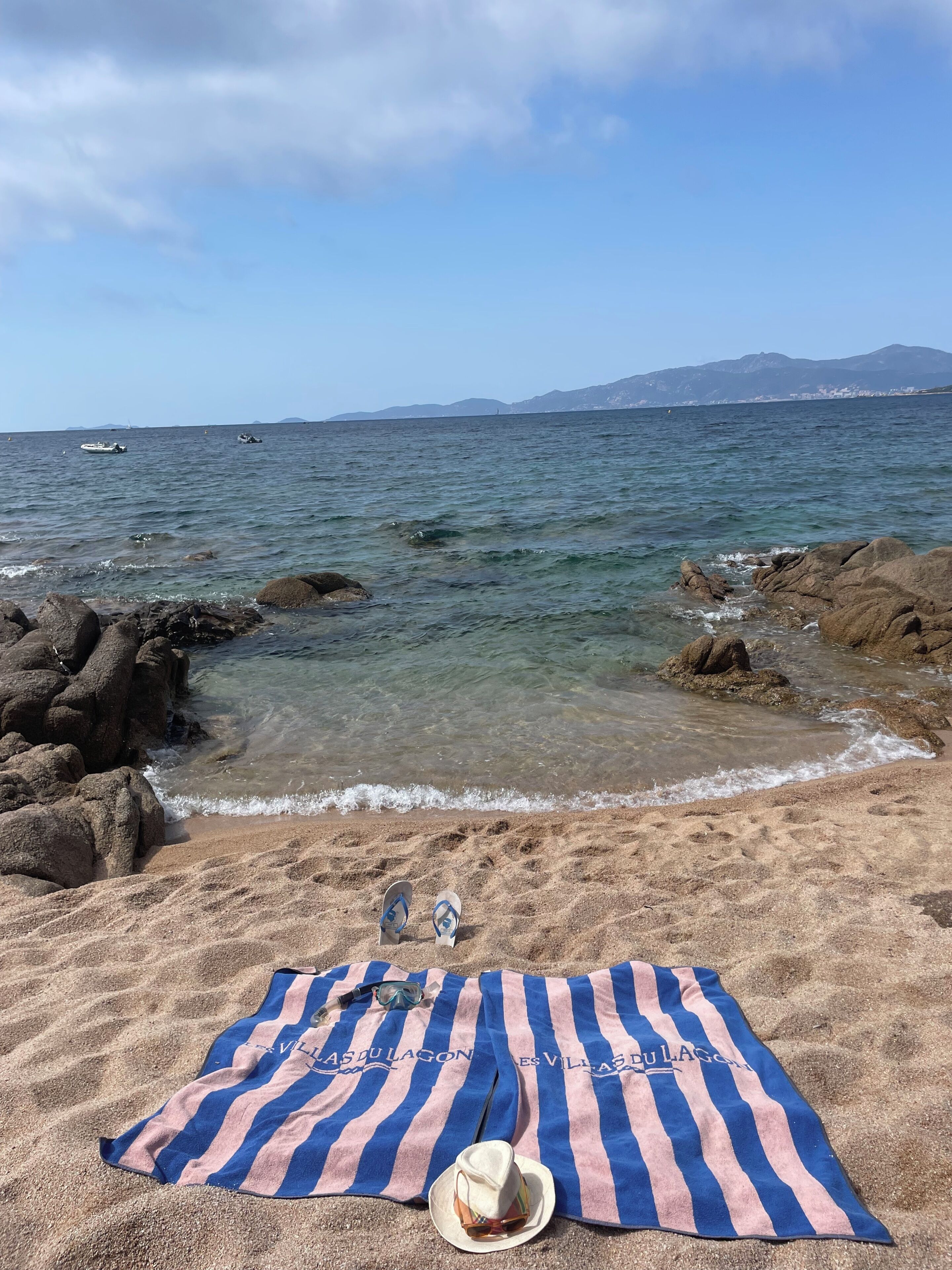A beach with a towel and hat on the sand, a rocky shoreline, and calm sea.