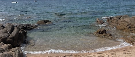 A beach with a towel and hat on the sand, a rocky shoreline, and calm sea.