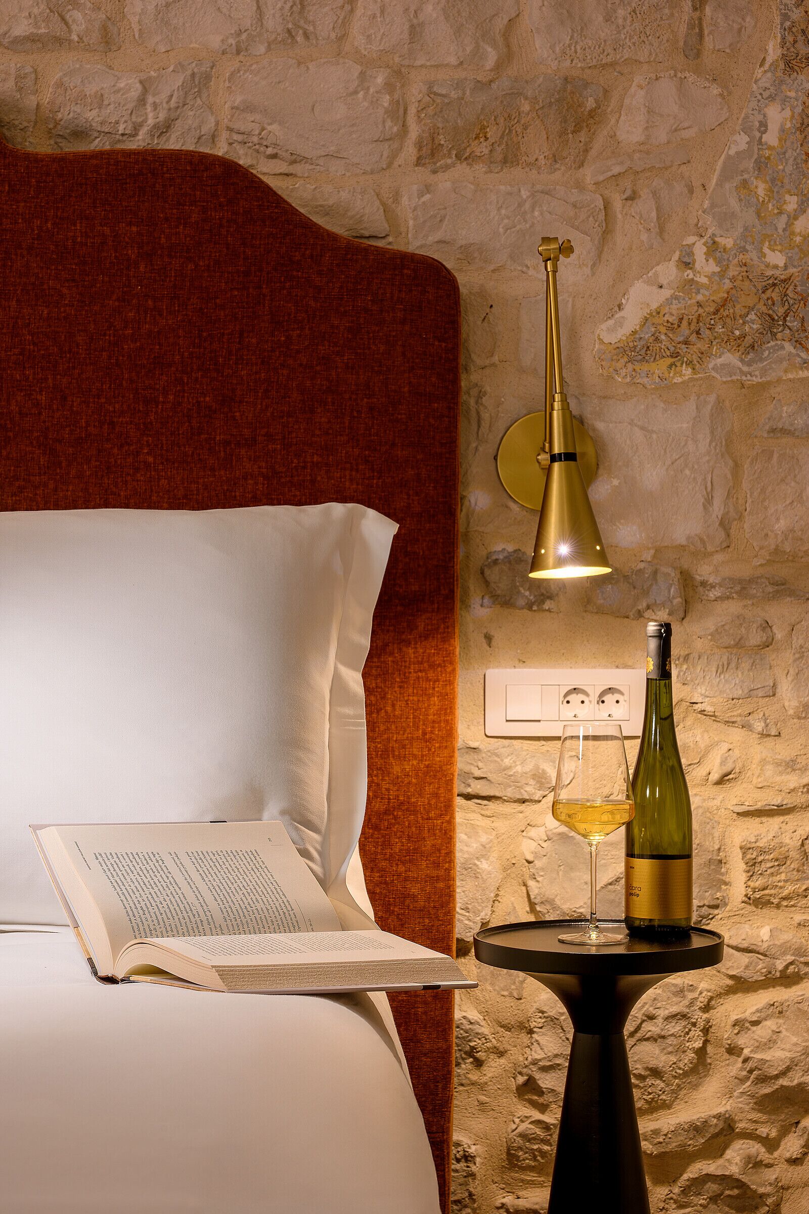 A bed with a white pillow and an open book, a small round table with a wine bottle and glass, and a wall-mounted lamp.