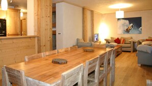 A wooden dining table with chairs, a kitchen area with wooden cabinets, a living room with a sofa and a painting, and a ceiling with pendant lights. - Alpine Apartment With Beautiful Views (Les Deux Alpes)