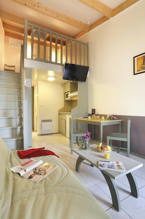 Una habitación con sofá, una mesa con periódico, una pequeña cocina y un televisor montado en la pared.