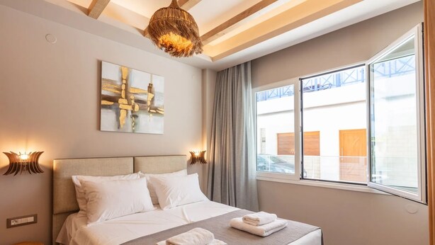 A hotel room with a bed, a window, a painting, and a ceiling light fixture. - Modern Luxury 2-Bed Apt, in the heart of Stalis and steps from the Beach. (Heraklion)