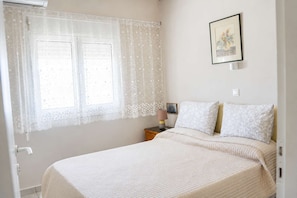 Un dormitorio con cama, mesita de noche, lámpara, un cuadro enmarcado en la pared y ventana con cortinas translúcidas.
