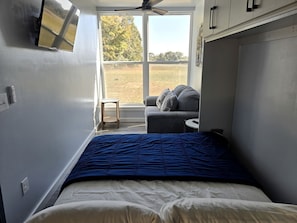 A bedroom with a bed, a small table, a chair, and a window with a view of a field.