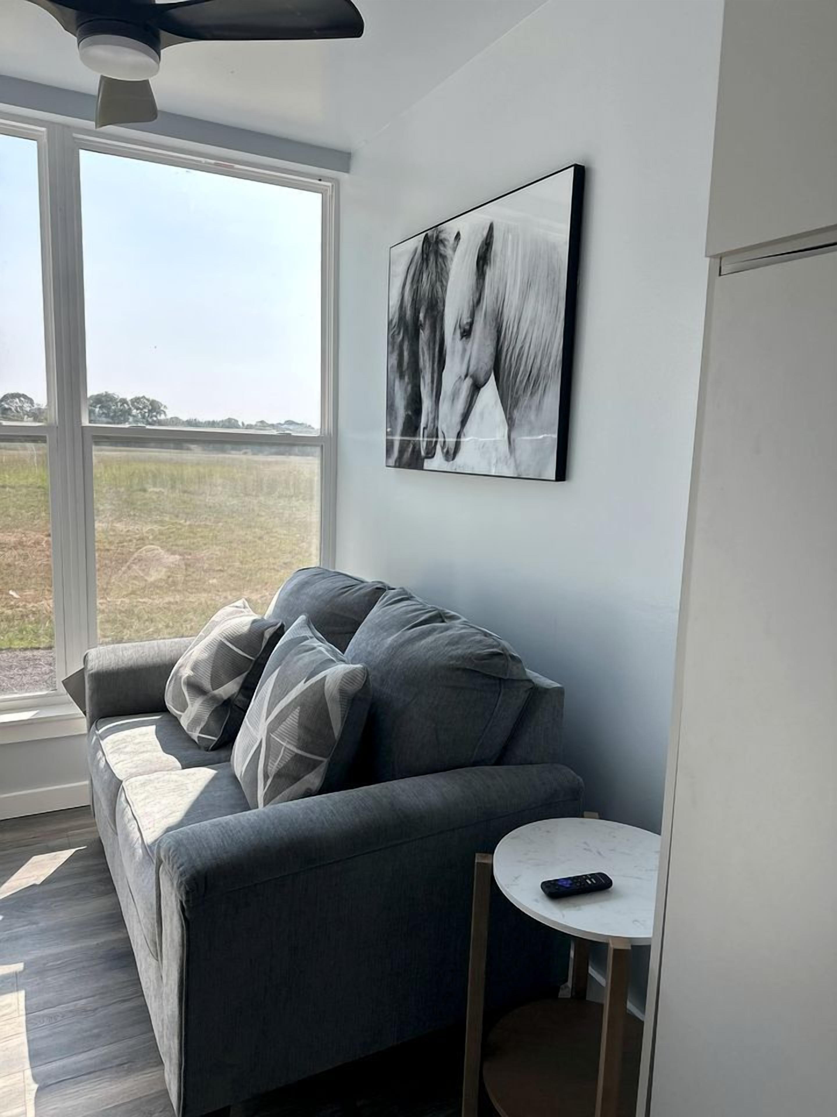 A modern living room with a grey sofa, a small white table, and a large window with a view of a field.