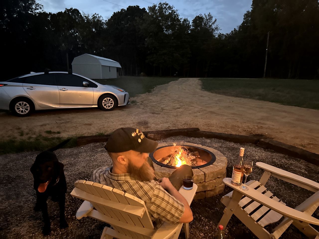 Une personne assise près d’un feu de camp, avec un chien, une voiture et une remorque en arrière-plan.