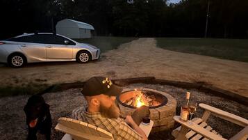 Une personne assise près d’un feu de camp, avec un chien, une voiture et une remorque en arrière-plan.