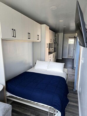 Een kleine slaapkamer met een bed, witte kasten, een televisie en een kitchenette.