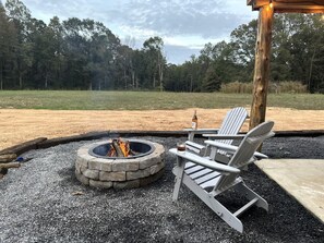 En eldstad med två Adirondack-stolar, en flaska och ett bord av betong, placerat på en grusbelagd yta med en skog i bakgrunden.