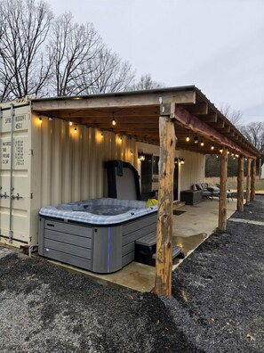 Ett bubbelbad under en pergola av trä, dekorerat med ljusslingor, ett skyddat sittområde och en konstruktion byggd av en fraktcontainer.