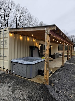 A hot tub under a wooden pergola with string lights, a covered seating area, and a shipping container structure.