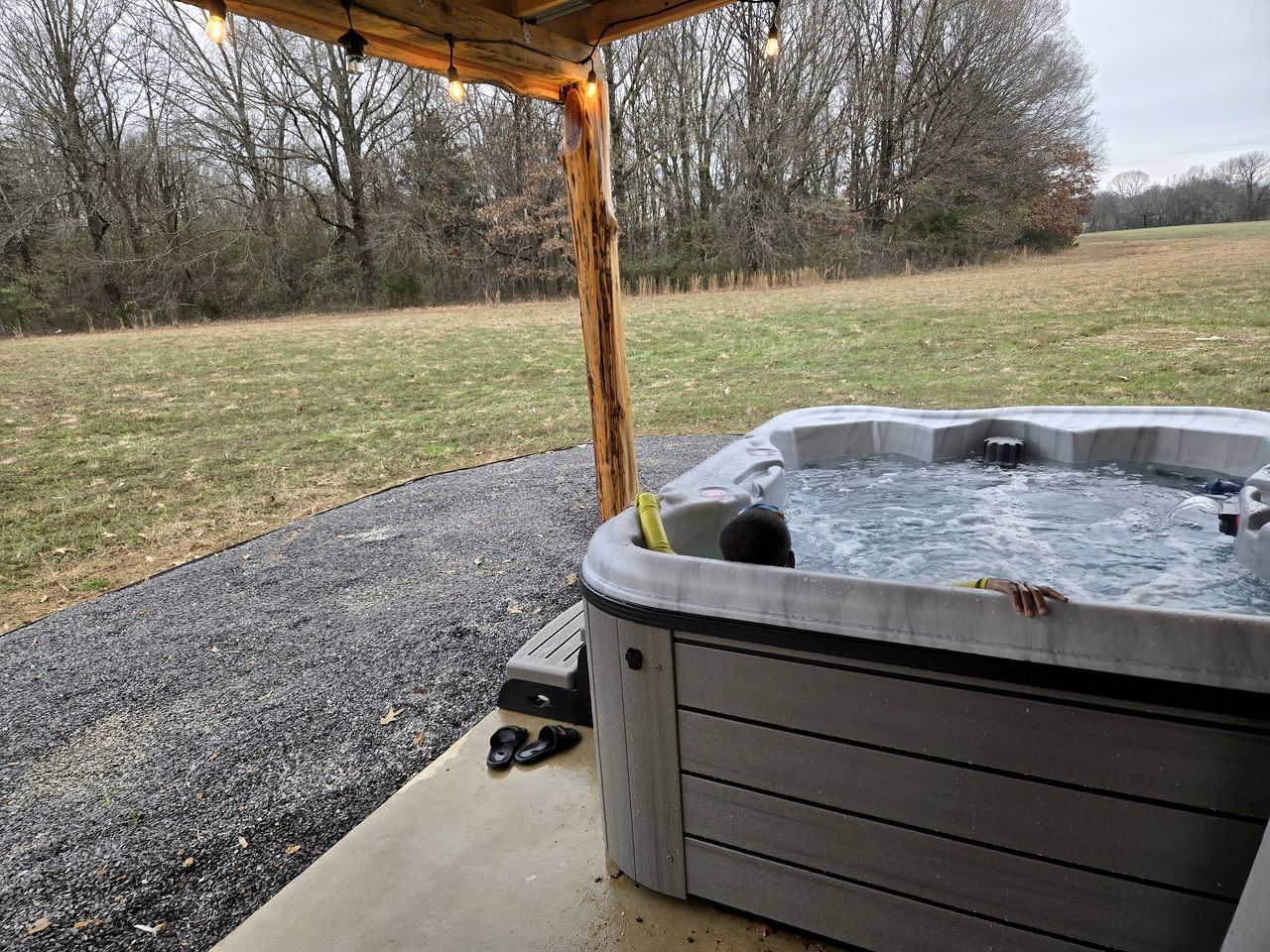 Un bain à remous extérieur, surmonté d’une pergola en bois, entouré d’un champ verdoyant et d’arbres.