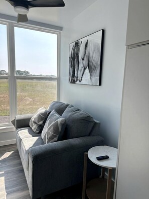 A modern living room with a grey sofa, a small white table, and a large window with a view of a field.