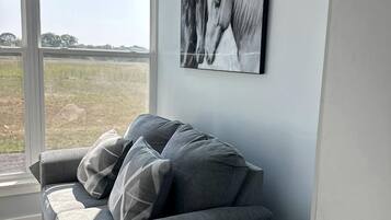 A modern living room with a grey sofa, a small white table, and a large window with a view of a field.