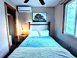 Een slaapkamer met een bed, een nachtkastje, een wandairconditioner, een ingelijste foto en een raam.