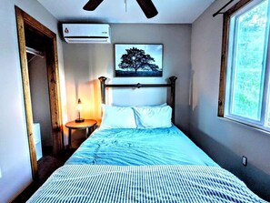 Un dormitorio con cama, mesita de noche, aire acondicionado de pared, un cuadro enmarcado y una ventana.