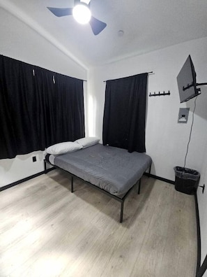 A single bed with a grey mattress, black curtains, a ceiling fan, and a small trash bin. - Peaceful Tiny Cabin Escape With Onsite Play Area Close To River Adventures in Houston, Alaska (Houston)
