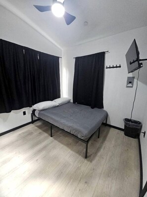 A single bed with a grey mattress, black curtains, a ceiling fan, and a small trash bin. - Peaceful Tiny Cabin Escape With Onsite Play Area Close To River Adventures in Houston, Alaska (Houston)