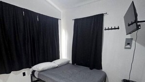 A single bed with a grey mattress, black curtains, a ceiling fan, and a small trash bin.