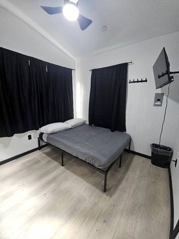 A single bed with a grey mattress, black curtains, a ceiling fan, and a small trash bin.