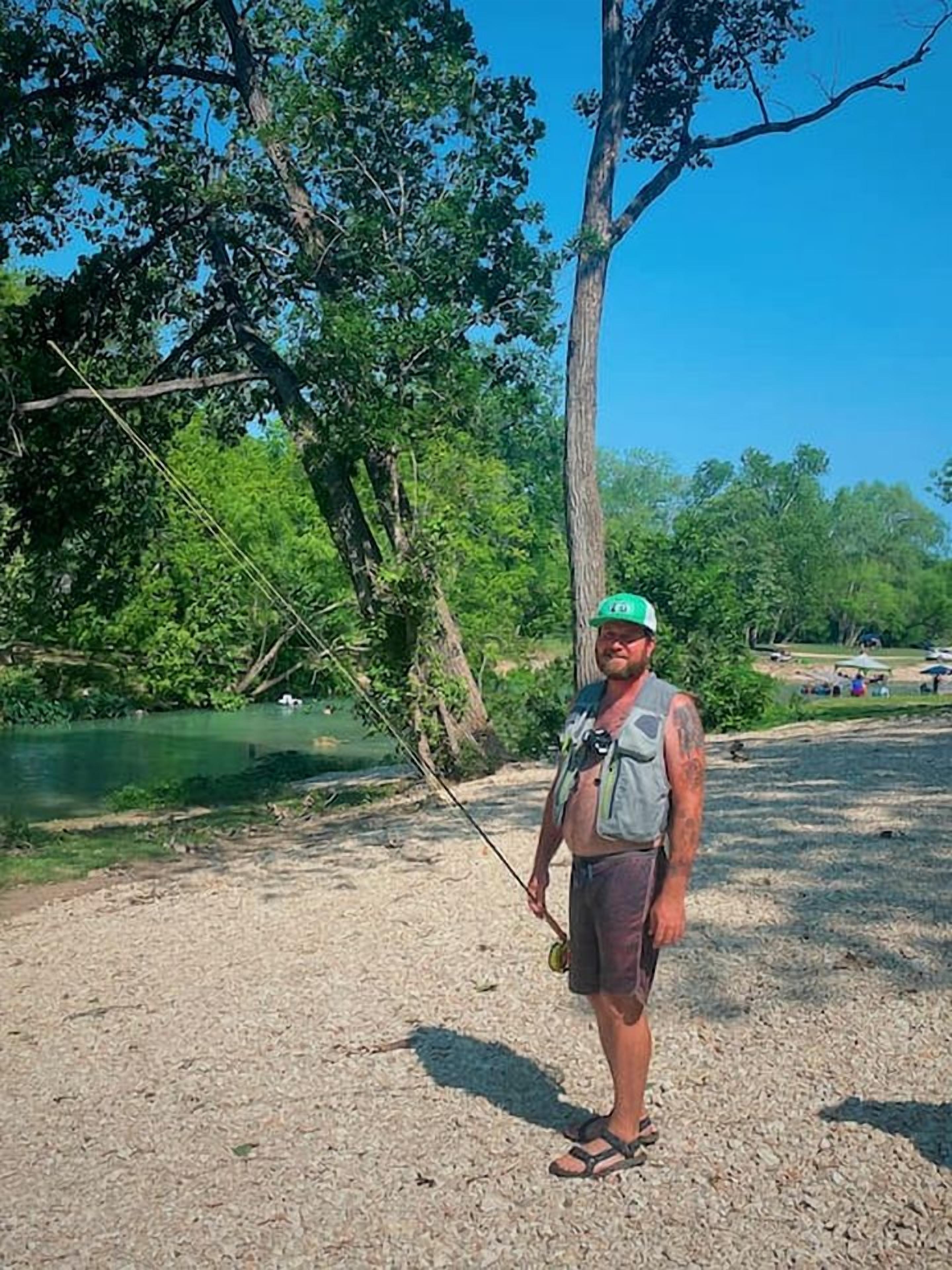 Une personne se tient à l’extérieur, tenant une canne à pêche, avec des arbres et une étendue d’eau en arrière-plan.