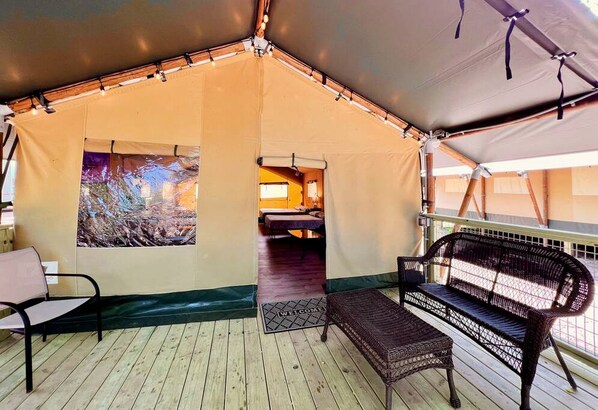 Alojamiento con carpa, una terraza de madera, un banco, una cama y vistas a un bosque.
