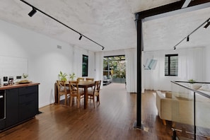 A modern kitchen with wooden flooring, a dining area with a table and chairs, and a glass railing overlooking a garden. - Sunlit 3-bed Coastal Stay With Private Deck (Sydney)