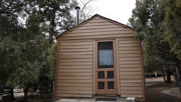 Une petite cabane en bois, d’un seul étage, avec une porte et une fenêtre, entourée d’arbres.
