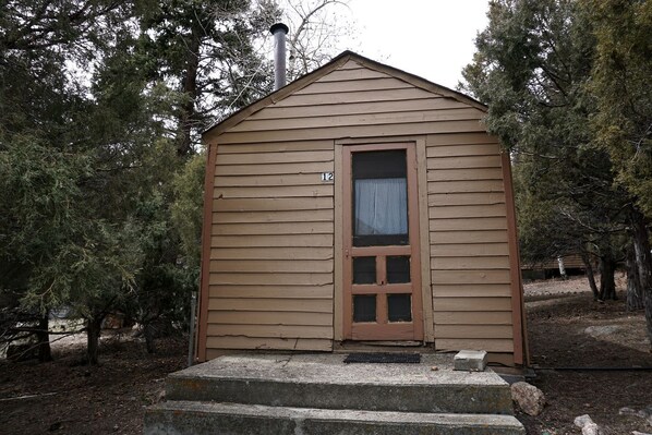 A small, single-story wooden cabin with a door and a window, surrounded by trees.