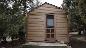 A small, single-story wooden cabin with a door and a window, surrounded by trees.