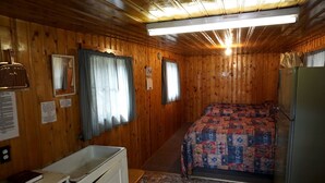 Une cabine comprenant un lit, une petite kitchenette, une fenêtre avec des rideaux et un ventilateur de plafond.
